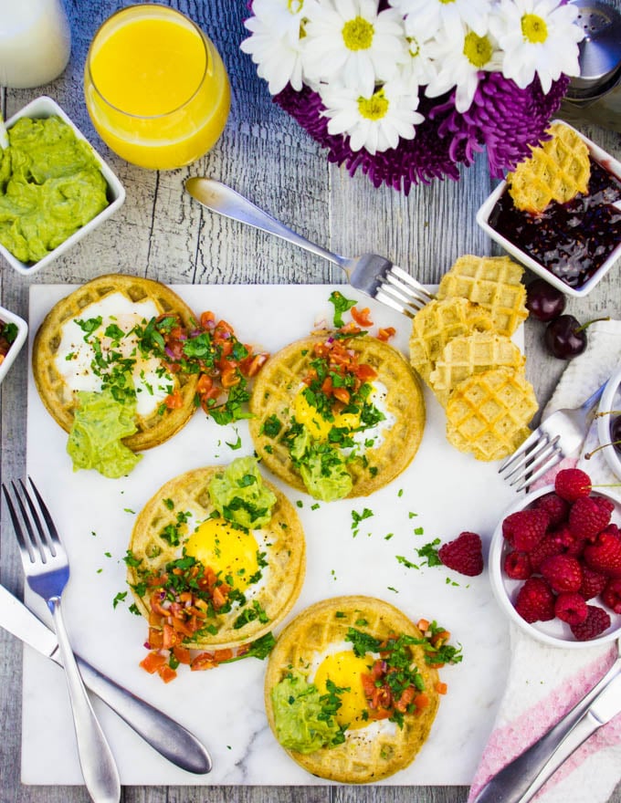 Finished breakfast table with four egg in a hole waffles, some flowers and berries, extra guacamole and jam on the side.