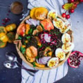 A top view of a kale salad bowl with fruits, hard boiled eggs, spicy lentils surrounded by fresh fruits, a kitchen towel and some spoons.