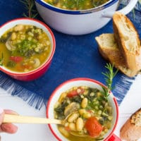 A spoon holding some soup showing white beans, chunky tomatoes and kale over a bowl of soup