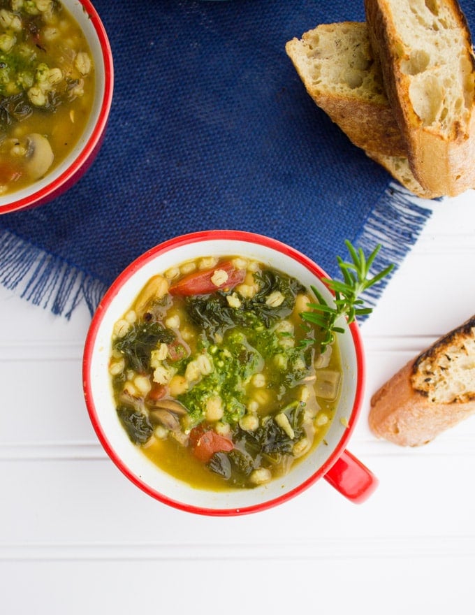 A top view of a white bean soup with kale, some pesto and a sprig of rosemary and some crunchy bread.