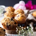 A bran muffin over a wooden board with more bran muffins at the back surrounded by small white flowers