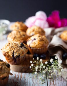 A bran muffin over a wooden board with more bran muffins at the back surrounded by small white flowers