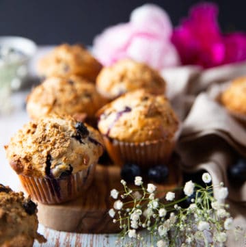 A bran muffin over a wooden board with more bran muffins at the back surrounded by small white flowers