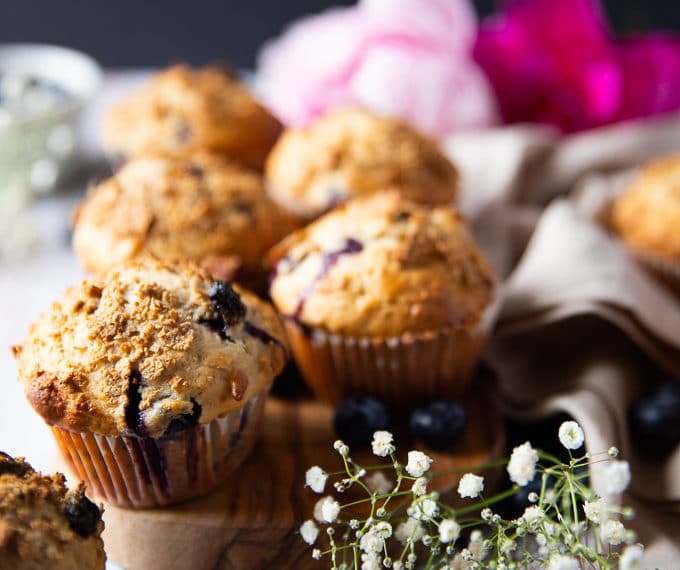 A bran muffin over a wooden board with more bran muffins at the back surrounded by small white flowers