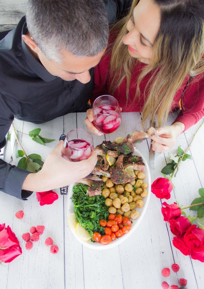 Two people lookig at each other and enjoying the lamb platter together for Valentine's Day. One of them holding a lamb chop while the other holding the drink.