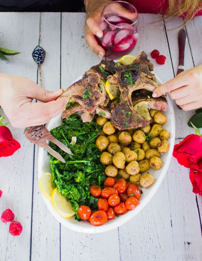 two hands holding a lamb chop each over the lamb chops platter with potatoes, broccoli, and tomatoes.