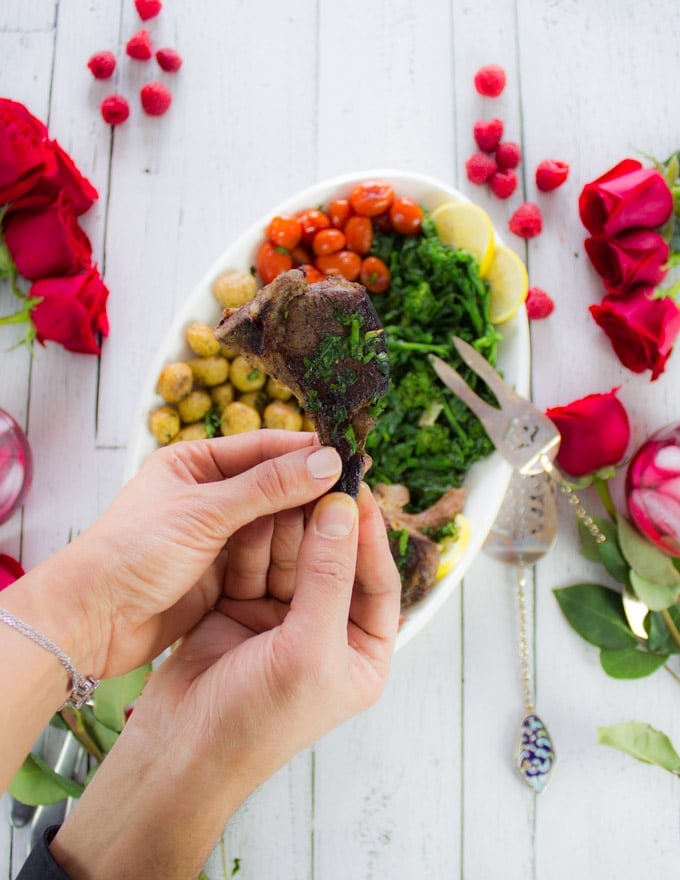 two hands sharing and holding a single lamb chop over a platter of lamb chops and roses, raspberries and drinks for a date night table set up