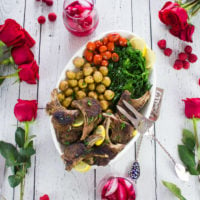 A full platter of lamb chops, roast potatoes, garlic broccolini and tomatoes, surrounded by red roses and drinks