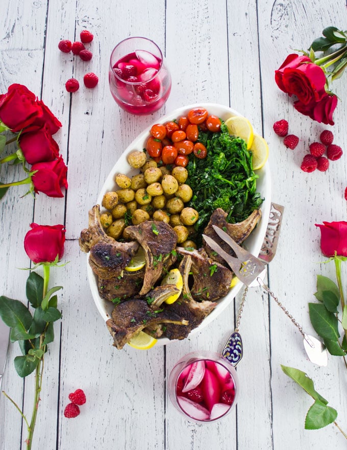 A full platter of lamb chops, roast potatoes, garlic broccolini and tomatoes, surrounded by red roses and drinks