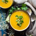 A bowl of butternut squash recipe surrounded by two spoons, some lime and cilantro.