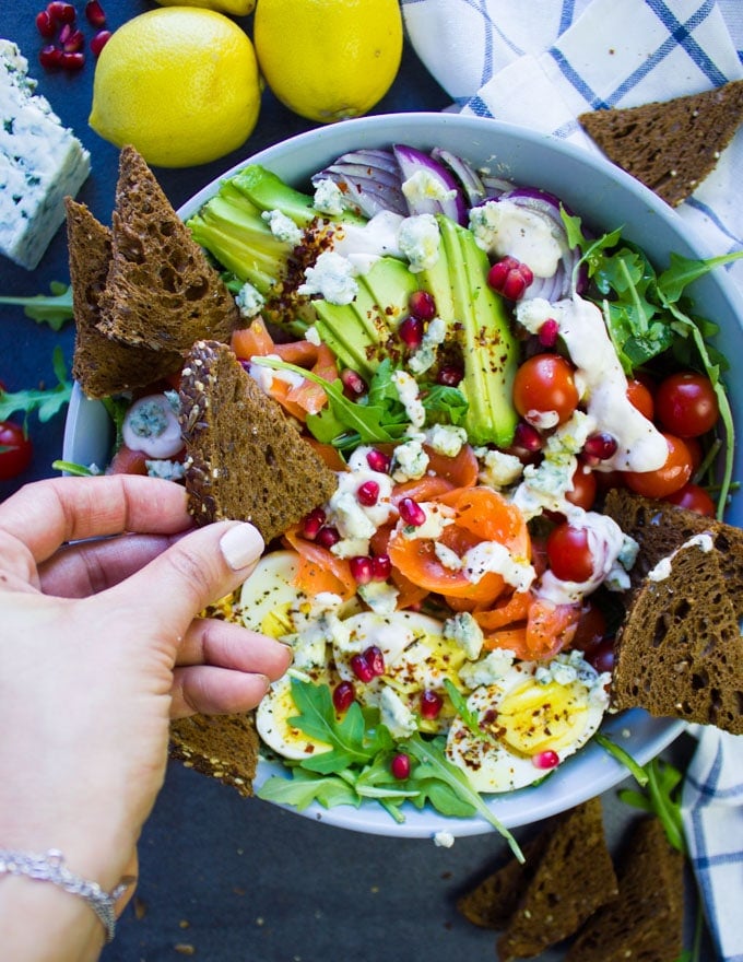 A hand holding a pumpernickel toast and digging in a bowl os smoked salmon salad with blue cheese dressing.