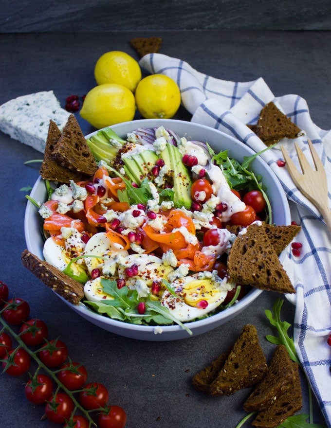 A bowl of smoked salmon salad and blue cheese dressing surrounded by a tea towel, cherry tomatoes and pumpernickel croutons.