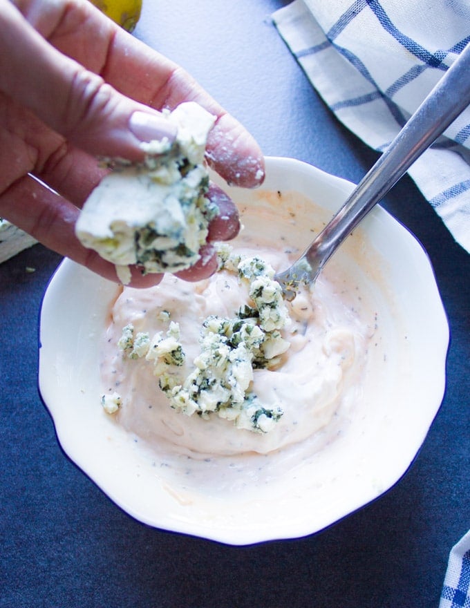 A hand holding blue cheese and crumbling it over a bowl of blue cheese dressing