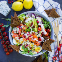 A huge bowl os smoked salmon salad with pumpernickel croutons, surrounded by a tea towel, some fresh lemons and two salad spoons.
