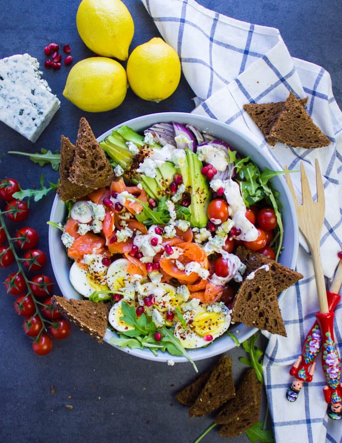 A huge bowl os smoked salmon salad with pumpernickel croutons, surrounded by a tea towel, some fresh lemons and two salad spoons.