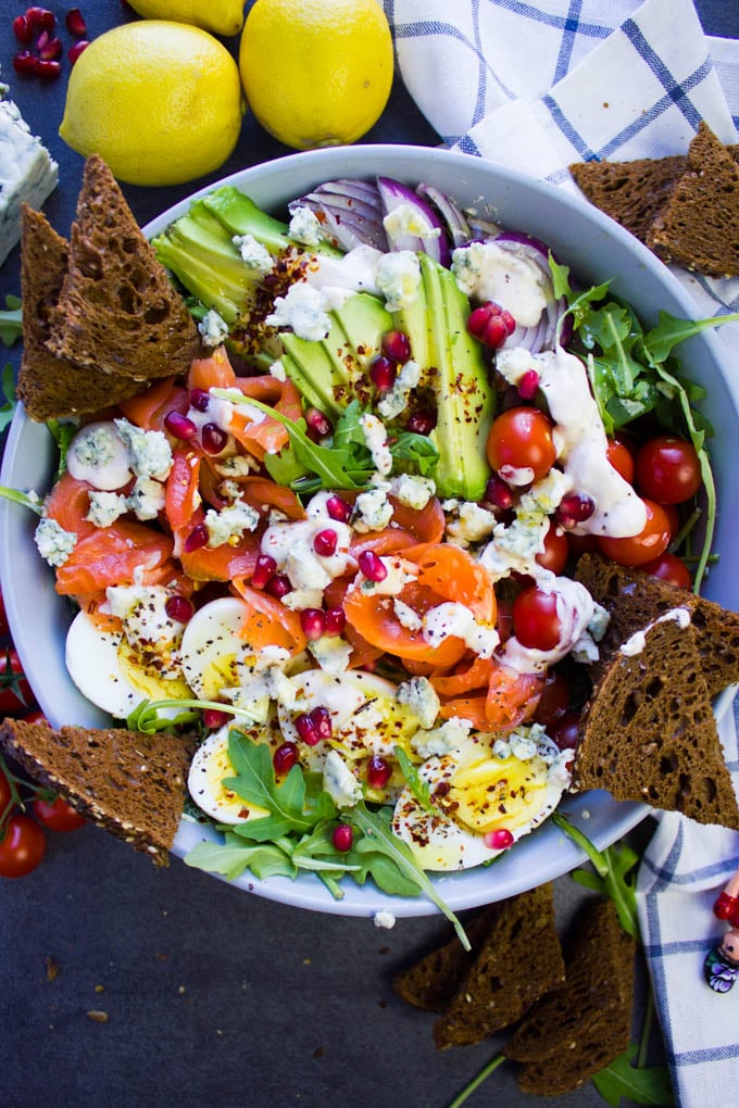 A huge bowl of smoked salmon salad with blue cheese dressing on top, pumpernickel croutons, sliced avocados, hard boiled egg and tomatoes.