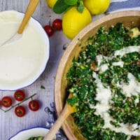 A half a bowl of tabouleh salad with lemons on a table and a bowl of tahini dressing