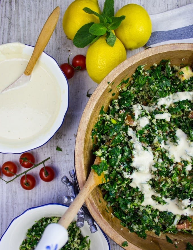 A half a bowl of tabouleh salad with lemons on a table and a bowl of tahini dressing 