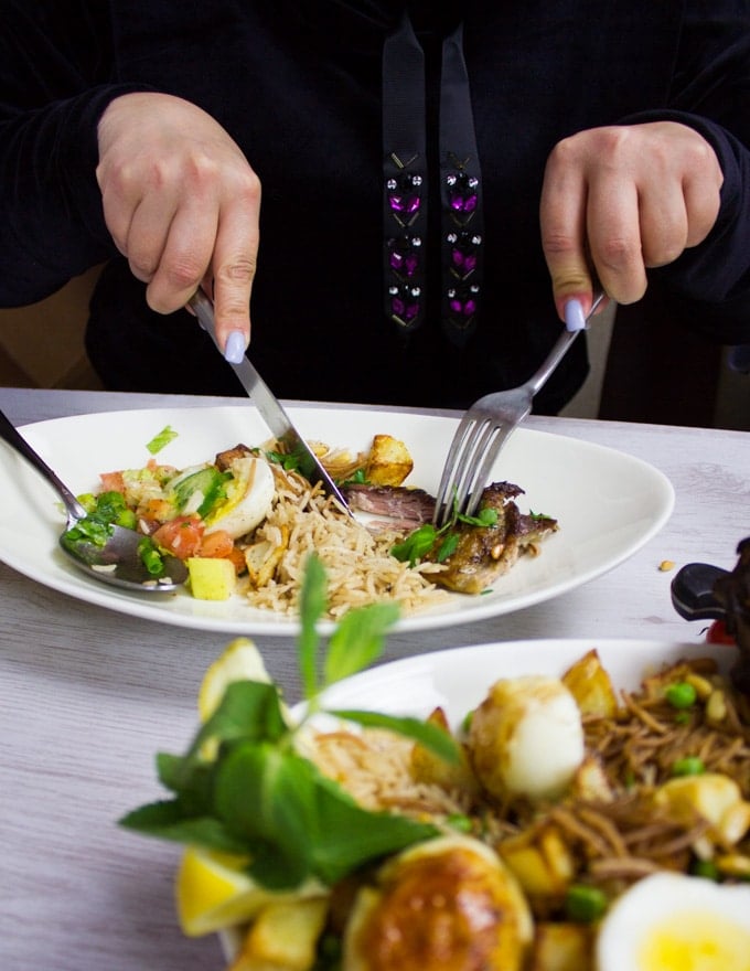 A plate with a bit of lamb shoulder, some rice and salad and a hand holding fork and knife eating and enjoying the meal