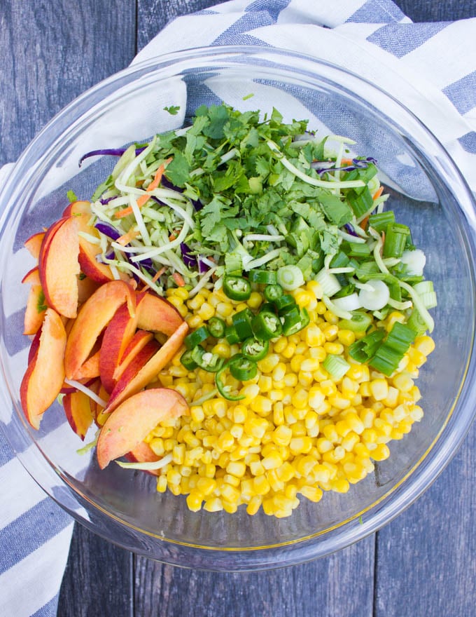 A big bowl of fish taco slaw ingredients including corn kernels, sliced peaches, broccoli slaw, jalapenos, scallions, cilantro