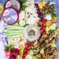 Lemon chicken and hummus platter showing hummus tube surrounded by lemon chicken, grilled lemon, olives, grilled tomatoes, charred bread, cucumbers, peach salad and feta cheese