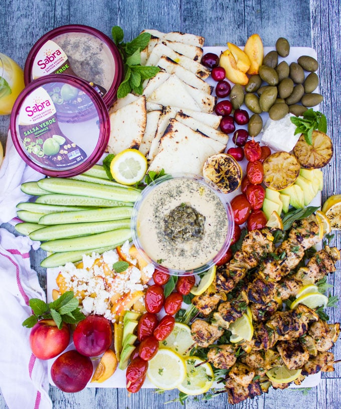 Lemon chicken and hummus platter showing hummus tube surrounded by lemon chicken, grilled lemon, olives, grilled tomatoes, charred bread, cucumbers, peach salad and feta cheese