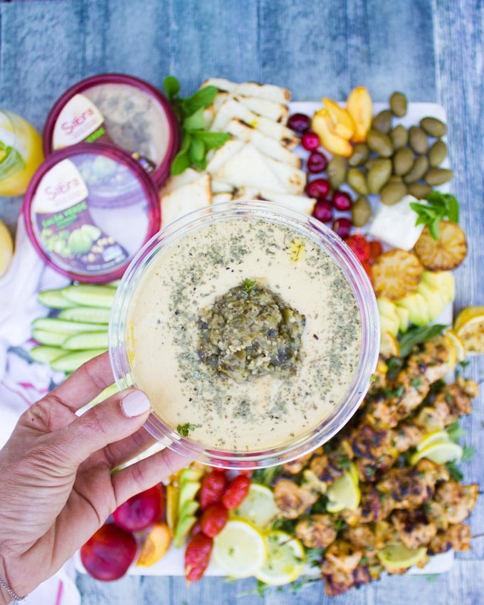 A hand holding a sabra hummus tub close up over the lemon chicken platter