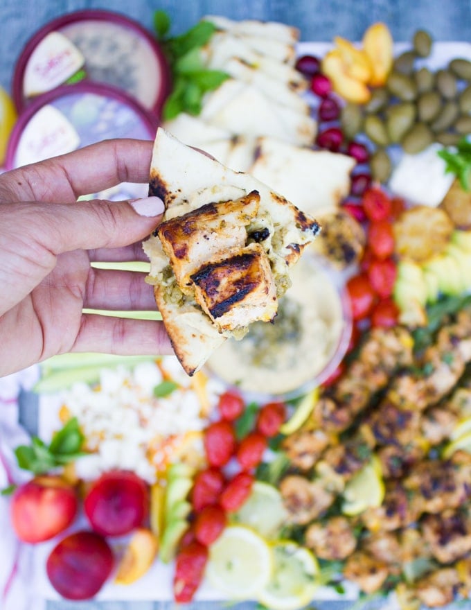 A hand holding a piece of charred bread spread with hummus and topped with a piece of lemon chicken