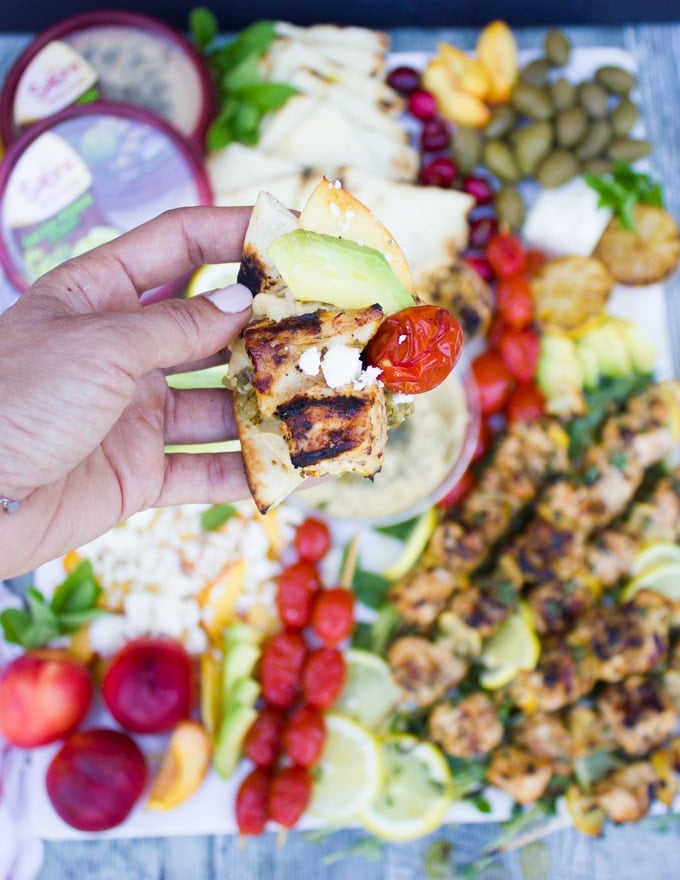 A hand holding a piece of bread topped with sabra hummus, lemon chicken and grilled tomatoes, sliced avocados and feta cheese