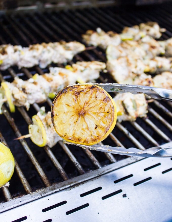 A tong showing a lemon grilled on the BBQ for the lemon chicken with the lemon chicken grilling behind