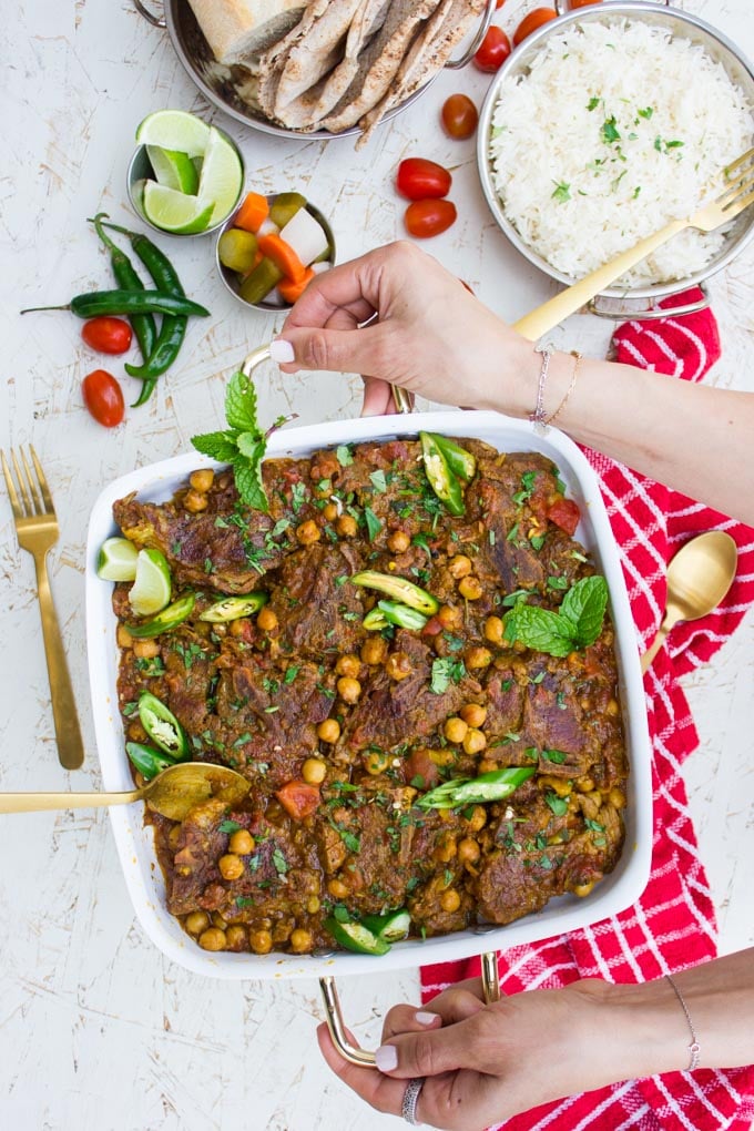 A hand holding a plate of roast lamb and chickpeas surrounded by a bowl of white rice, a red tea towel and some green chillies, fresh tomatoes