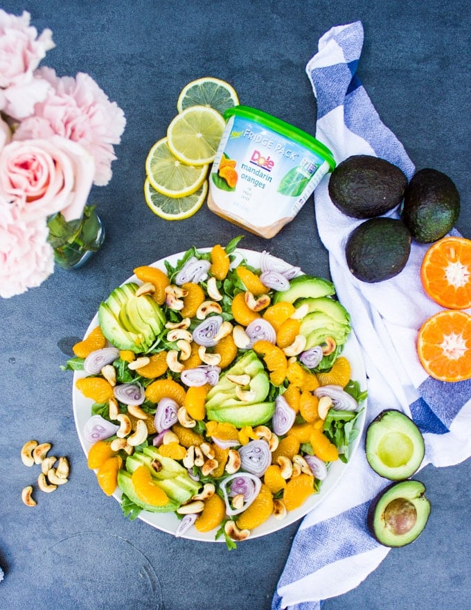 The finished avocado salad with citrus dressing, a vase with flowers, some whole avocados and some slices mandarin oranges around