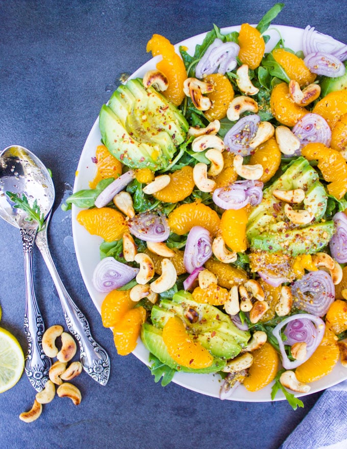 A half plate showing details of the Avocado Salad with oranges, citrus dressing, two spoons and some cashews