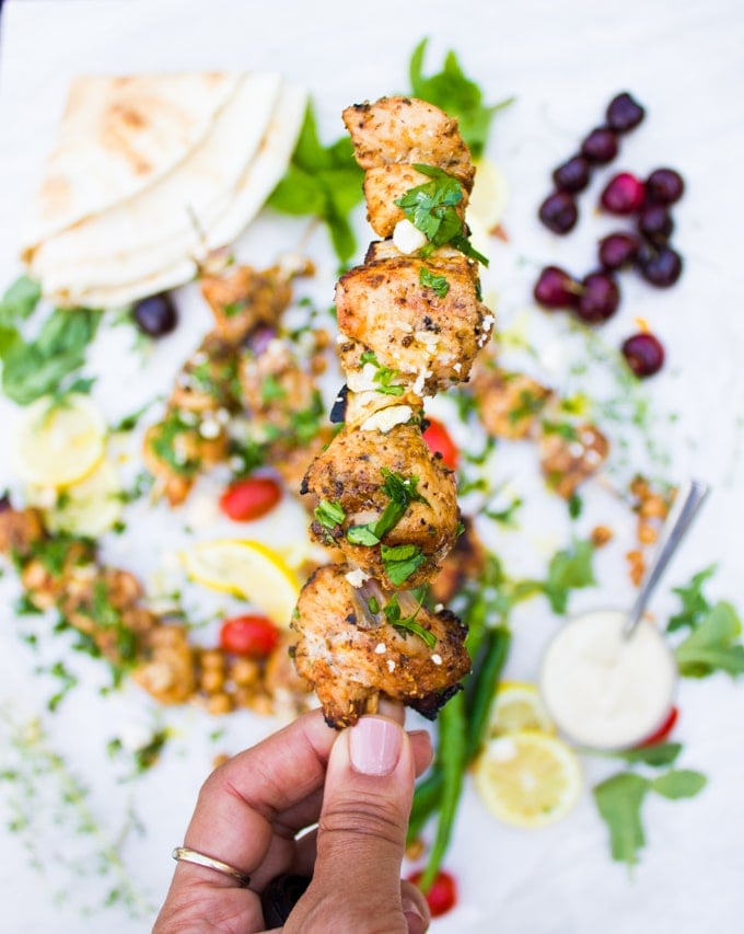 A hand holding a chicken skewer off from the grill, sprinkled with herbs and feta cheese