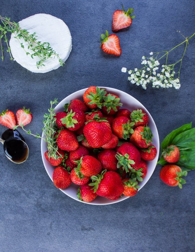 A bowl of Strawberries surrounded by a round of brie cheese, a spoonful of balsamic vinegar, fresh basil leaves and white flowers