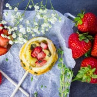 A savory hand pie on a lollipop stick with strawberries surrounded by fresh strawberries, fresh thyme and white flowers