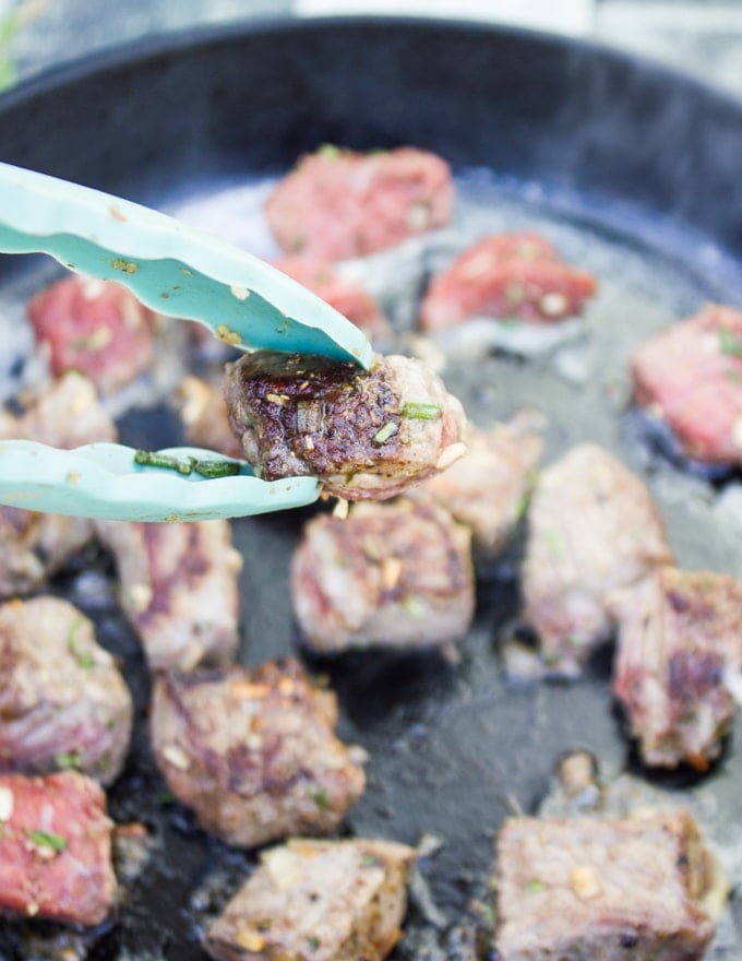 Steak Bites searing in a pan and a tong holding a piece of steak bite to show a close up of the searing 