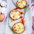 A series of three baked eggs in bread bowls surrounded by a basket of eggs and fresh baby tomatoes