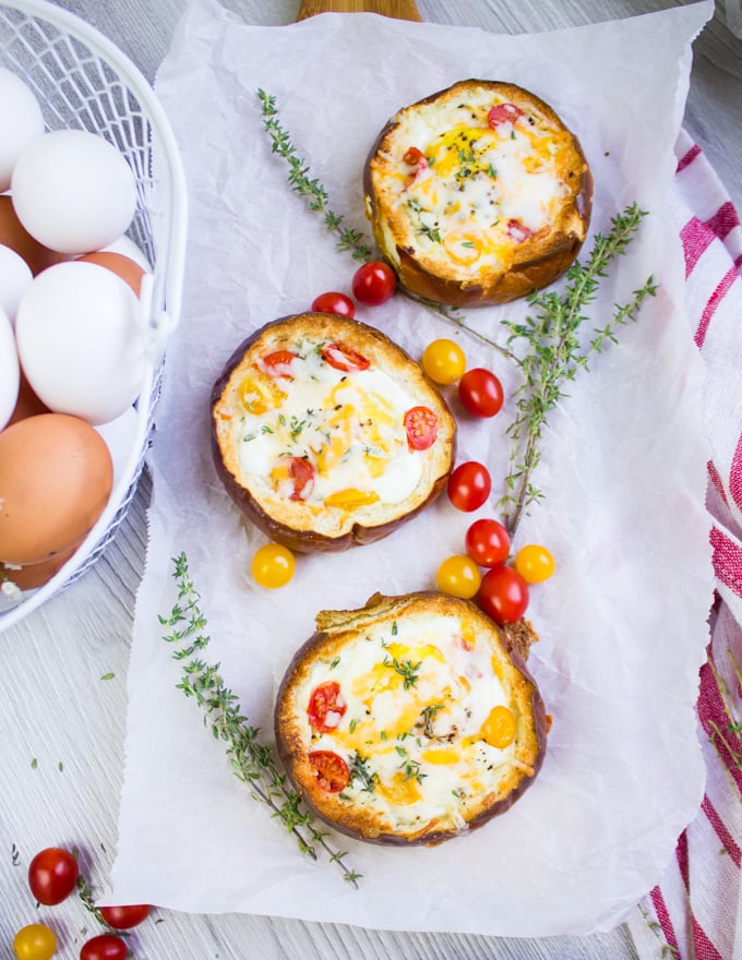 A series of three baked eggs in bread bowls surrounded by a basket of eggs and fresh baby tomatoes