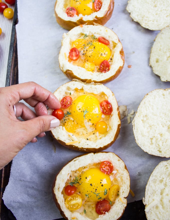 A hand adding diced tomatoes over the eggs in bread bowls