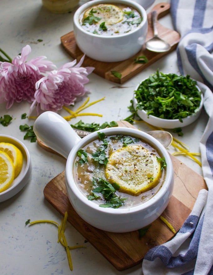 A close up of one bowl of soup topped with lemon slice, fresh herbs and surrounded by lemon slices and flowers