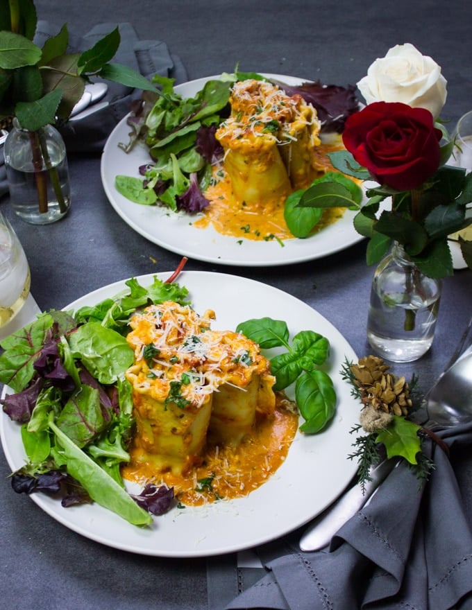 two plates with lasagna roll ups in rosa sauce on it. A red rose flower around that and a simple salad