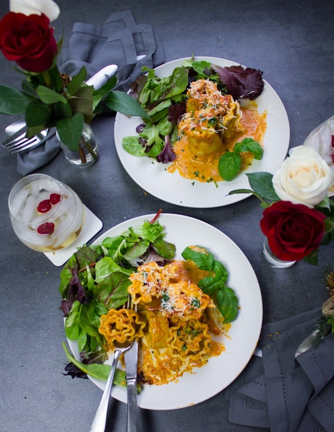 A fork and spoon eating off one plate of the lasagna roll ups and the other plate ready for serivng surrounded by red roses and drinks