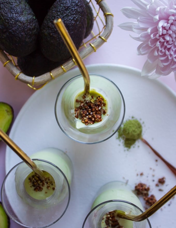 Three smoothie cups with straws and chocolate crumble on a white plate near a bowl of raw avocados 