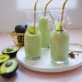 3 glasses of avocado smoothies with golden straws on a white plate