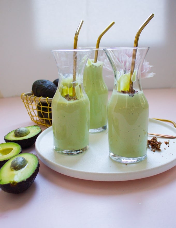 3 glasses of avocado smoothies with golden straws on a white plate