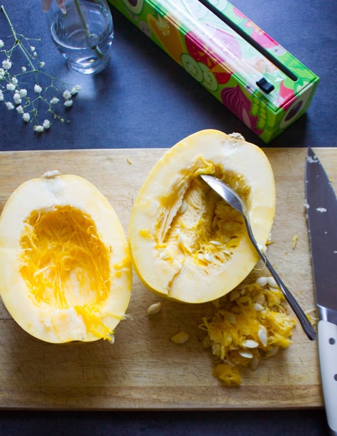 A spaghetti squash cut in half and a spoon removing the seeds from the inside on a cutting board