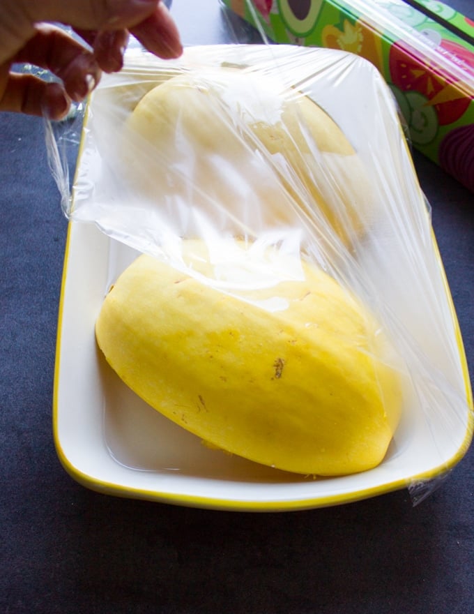 Two spaghetti squash halves placed on a dish and covered with plastic wrap ready to go in the microwave