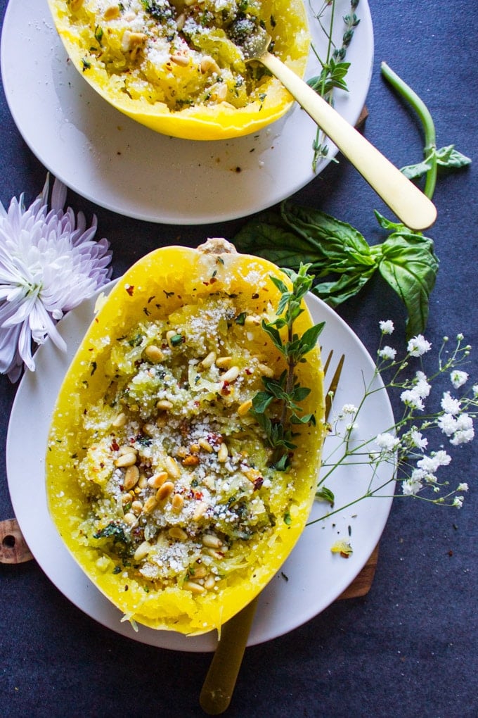 One cooked spaghetti squash recipe with parmesan, herbs, pine nuts and basil on a plate surrounded by a basil leaf and a flower.