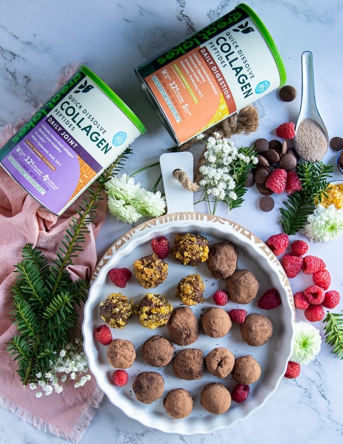 A plate of chocolate truffles and two boxes of collagen around it surrounded by flowers, raspberries and chocolate chunks